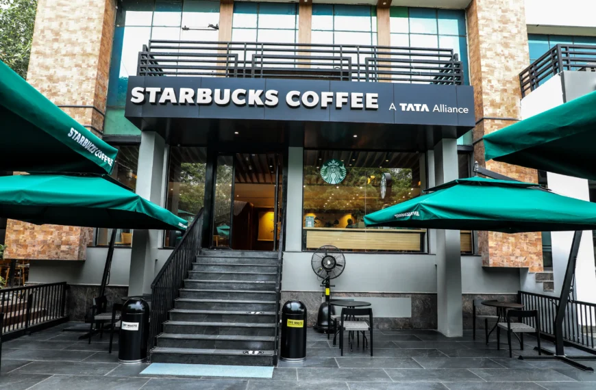 A Starbucks cafe exterior with green umbrellas and outdoor seating. The sign above the entrance reads "Starbucks Coffee, A Tata Alliance". This location offers a variety of drinks, including those listed on the Starbucks Menu With Calories.