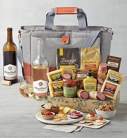  A picnic basket filled with gourmet food items, including wine, cheese, crackers, salami, and other snacks. The basket is open, showcasing the contents, and is accompanied by a gray tote bag for easy carrying.