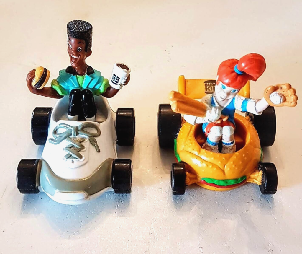 Two vintage Boomer
 Burger King Kids Club toys. The first toy is a white race car with a black figure driving it. The figure is holding a hamburger and a coffee cup. The second toy is a yellow race car shaped like a hamburger bun. The figure inside is holding a baseball bat and a baseball glove.