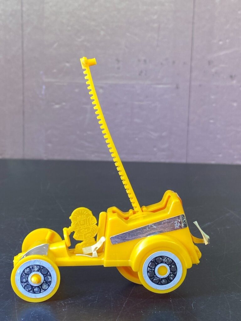 A yellow toy car with a long, retractable antenna. The car has four wheels with white tires and black detailing. There is a figure sitting in the driver's seat, and the car has a small spoiler on the back.